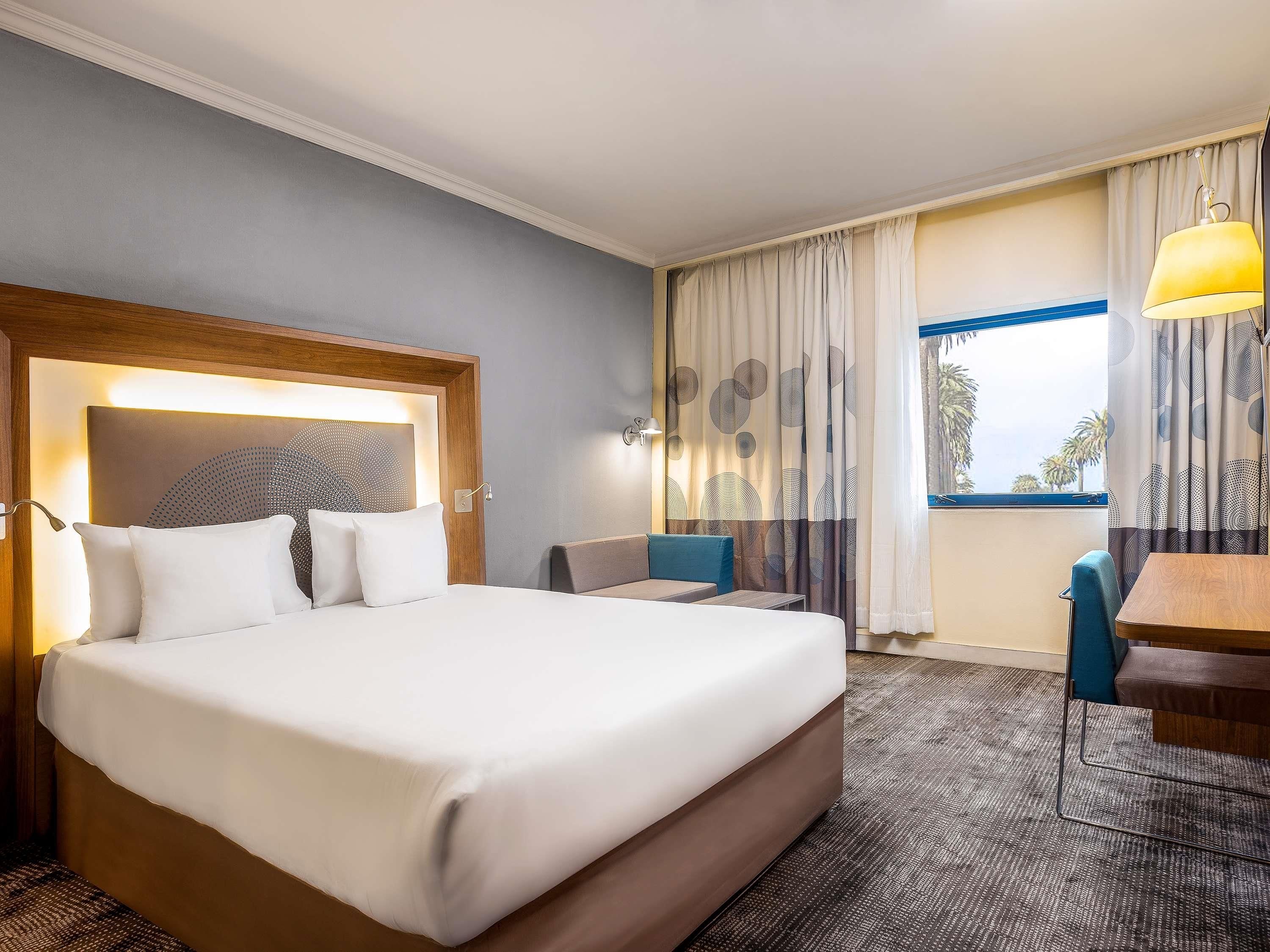 A hotel room with a large bed, a desk, a chair, and a window with curtains.