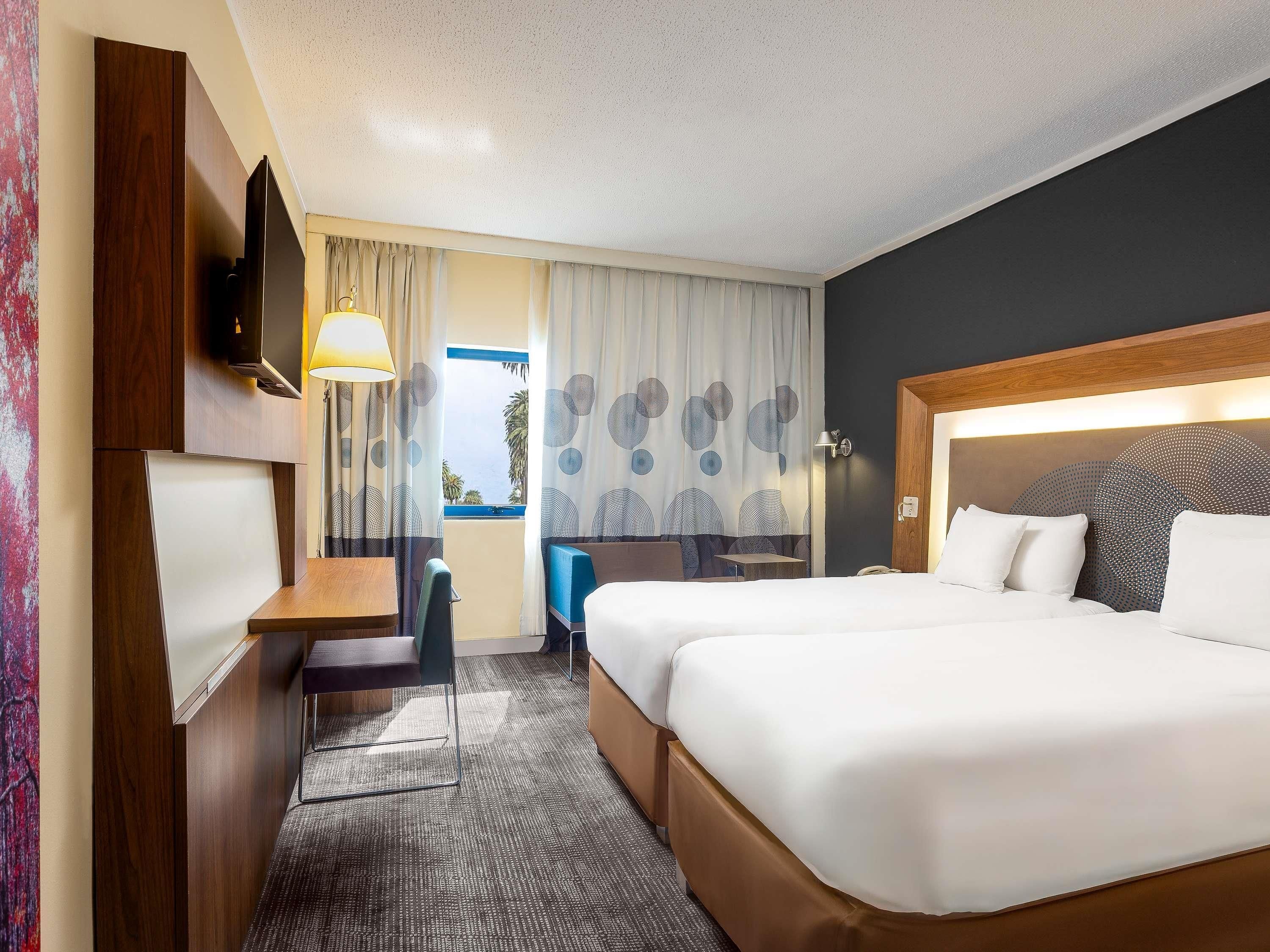 A hotel room with a bed, desk, chair, TV, and a view of the sea.