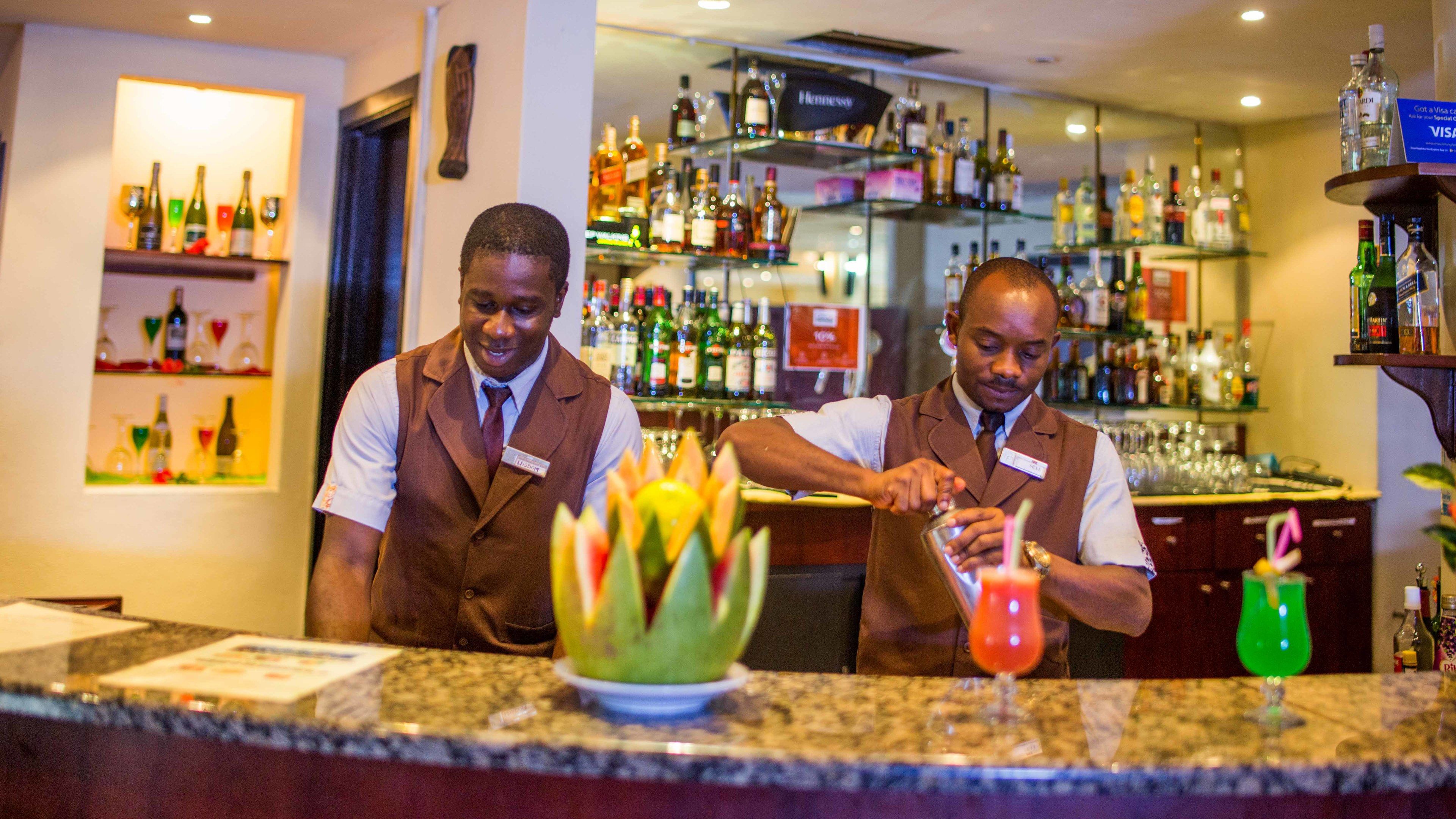 Deux barmen préparent des boissons au comptoir d’un bar, avec des étagères remplies de bouteilles de différents types d’alcool en arrière-plan.