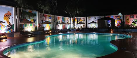 A nighttime swimming pool area with lounge chairs and umbrellas, surrounded by colorful murals.