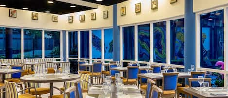 Un restaurante con varias mesas y sillas, amplias ventanas con obras de arte y un diseño moderno de techo.