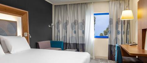 A hotel room with a bed, a desk, a chair, a lamp, and a view of palm trees through the window.