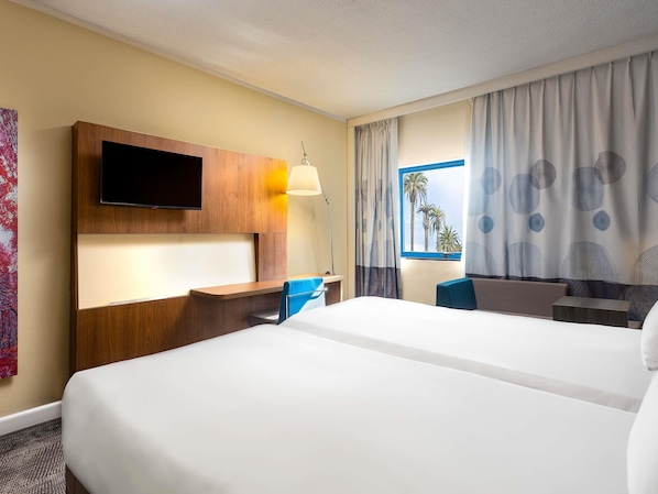 A hotel room with a large bed, a desk, a TV mounted on the wall, and a window with curtains.