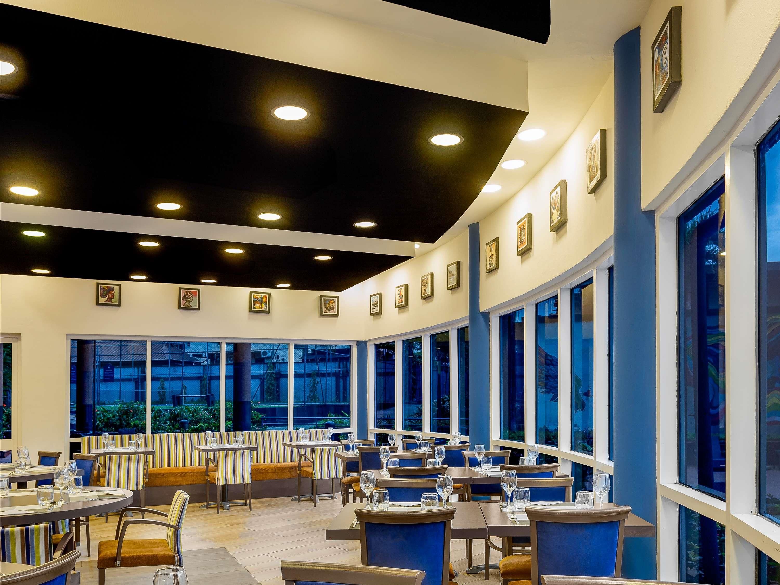 A restaurant with round tables and booths, large windows, and ceiling lights.