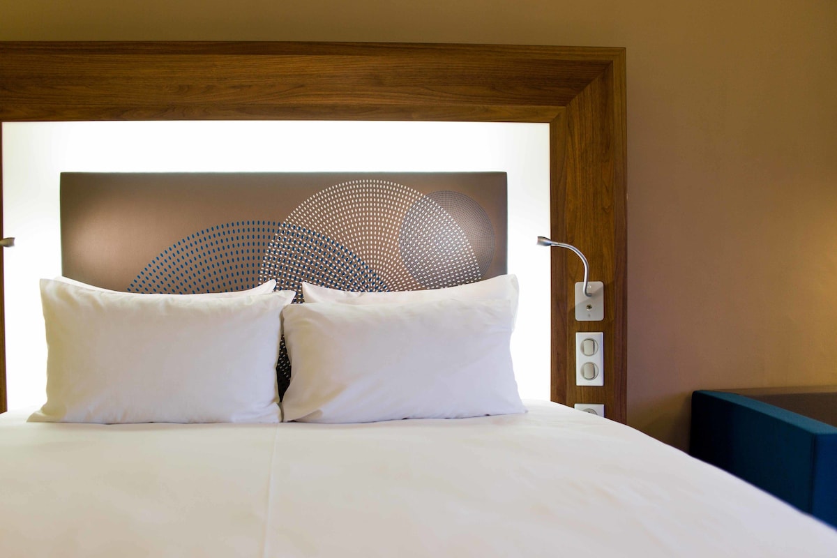 Habitación de hotel con una cama grande, dos almohadas blancas, un cabecero de madera y una lámpara fijada a la pared.