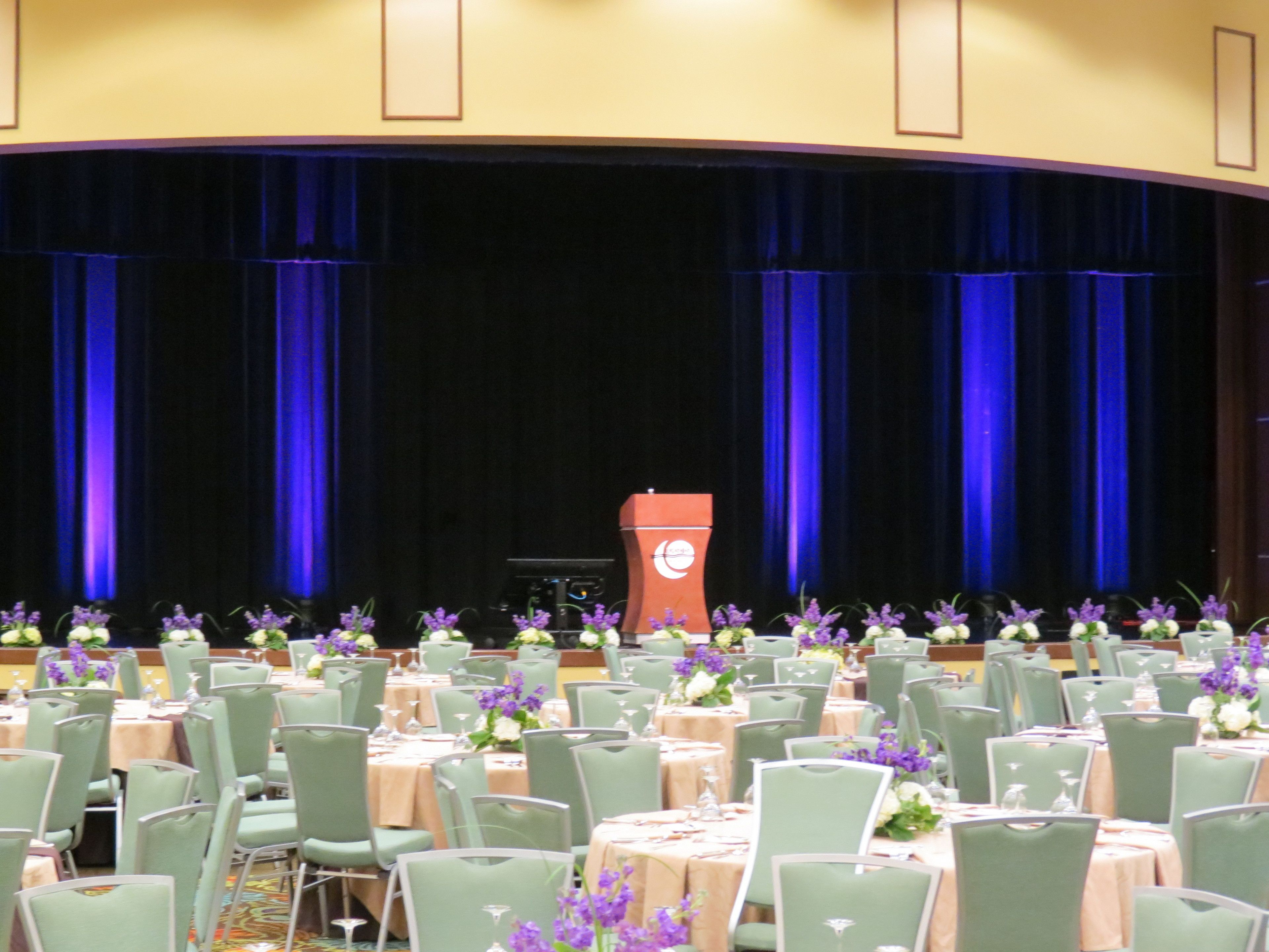 A banquet hall with rows of tables and chairs, a stage with a podium, and curtains.