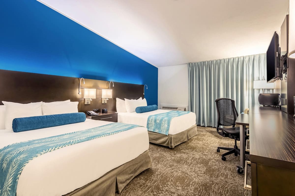 Habitación de hotel con dos camas, un escritorio, una silla, un televisor y un amplio ventanal con cortinas.