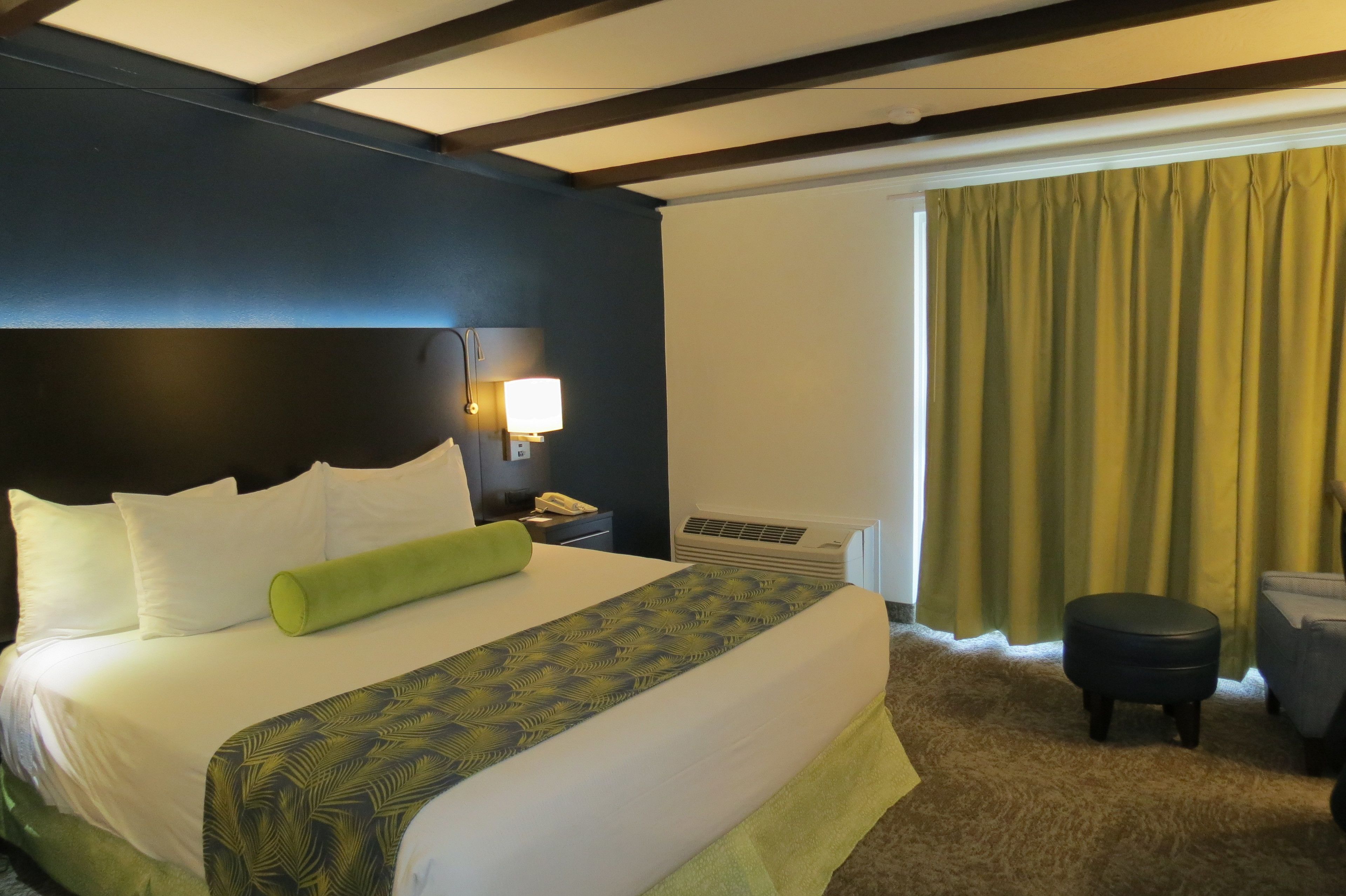 Ein Hotelzimmer mit einem großen Bett, einem Nachttisch mit Lampe, einem Fenster mit Vorhängen und einem kleinen Hocker.