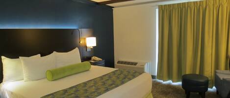 Ein Hotelzimmer mit einem großen Bett, einem Nachttisch mit Lampe, einem Fenster mit Vorhängen und einem kleinen Hocker.