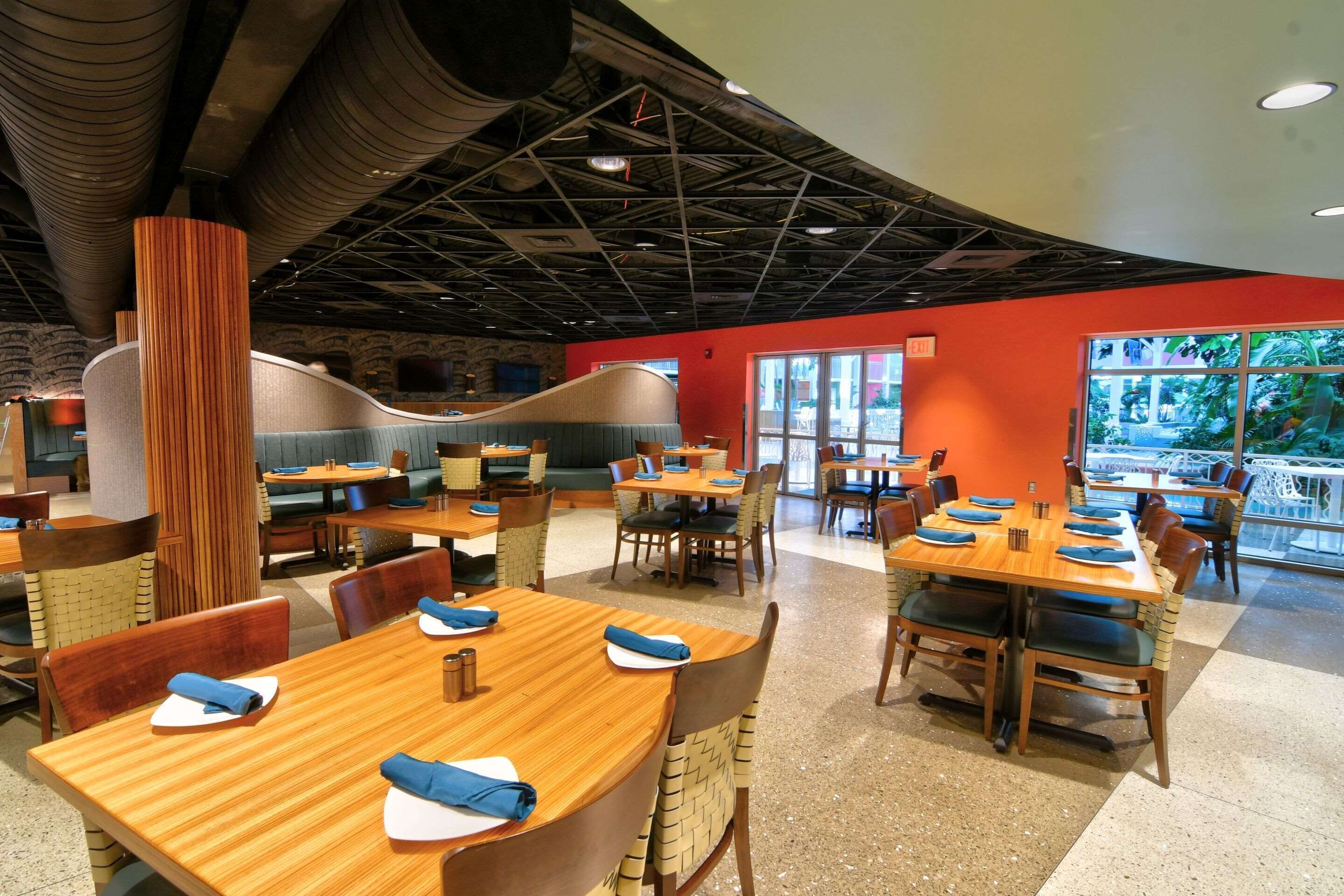 A spacious restaurant with multiple wooden tables and chairs, a high ceiling with exposed beams, and large windows offering a view of the outdoors.