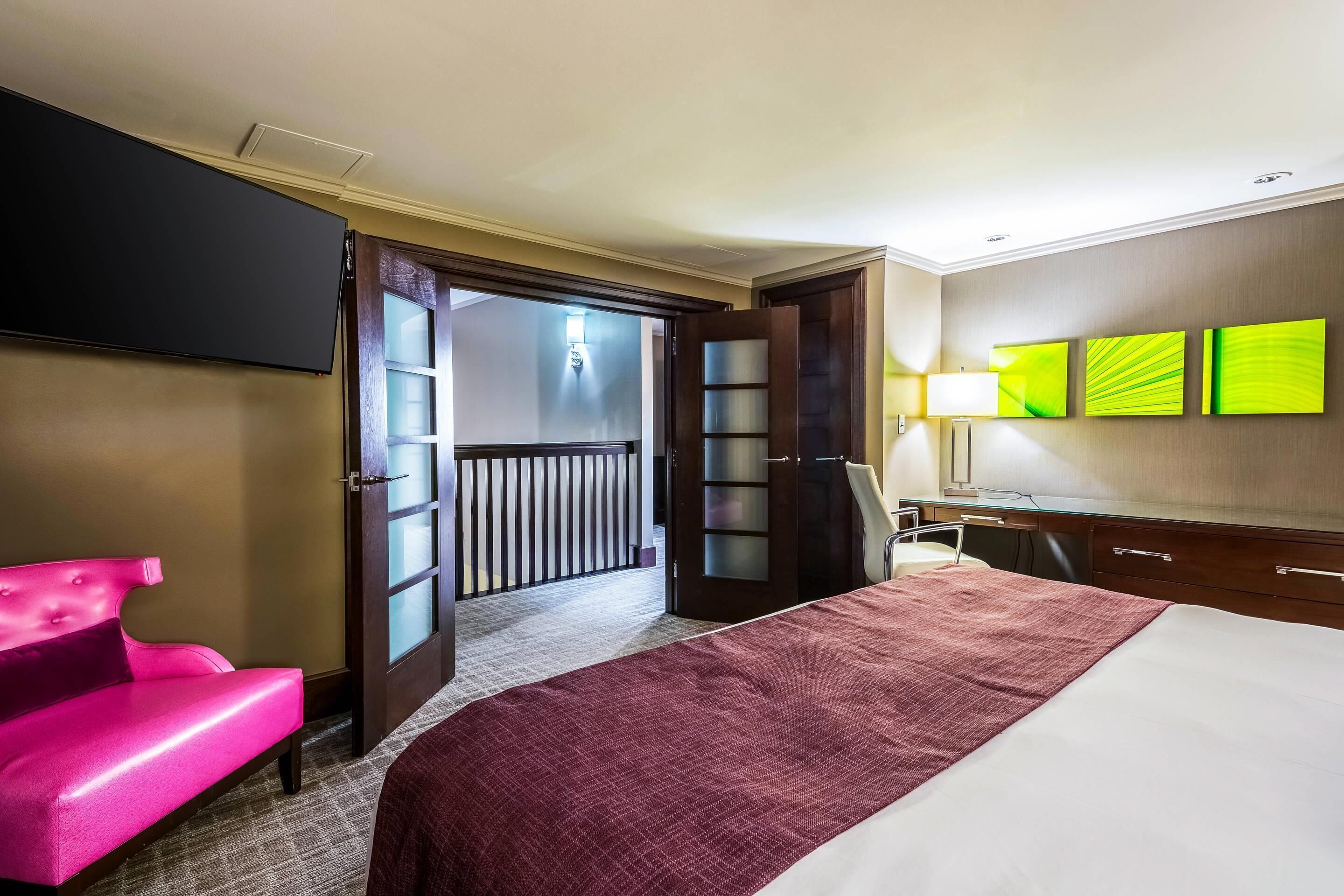 Une chambre d’hôtel avec un lit, une chaise rose, un bureau et un téléviseur.