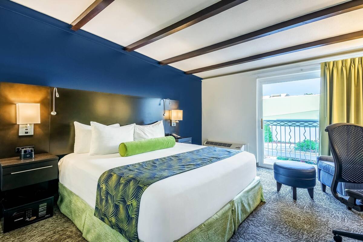 Habitación de hotel con una cama grande, dos lámparas de noche, un cojín verde, una ventana con vistas y un balcón con una silla.