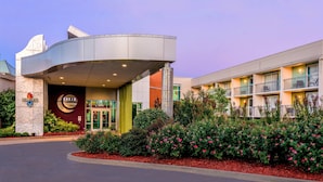 A hotel with a curved entrance, a prominent logo, and a landscaped garden with pink flowers. - Oasis Hotel & Conv. Center, an Ascend Collection Hotel (Springfield)