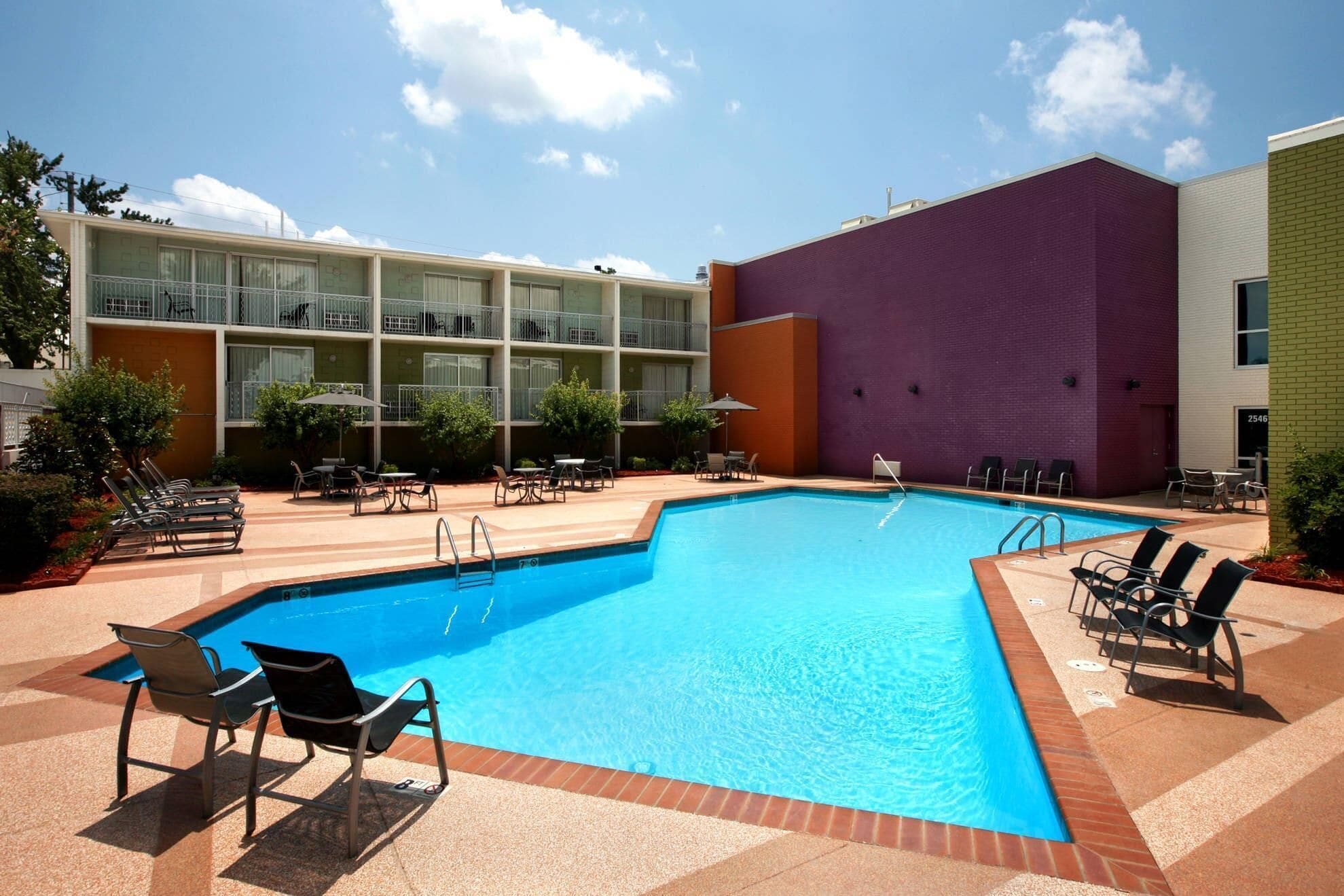 A swimming pool with lounge chairs and tables, surrounded by a building with multiple balconies and a brick wall.