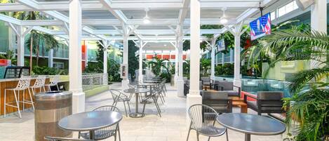 Un área de patio al aire libre con mesas y sillas, rodeada de vegetación y un cerramiento de vidrio.