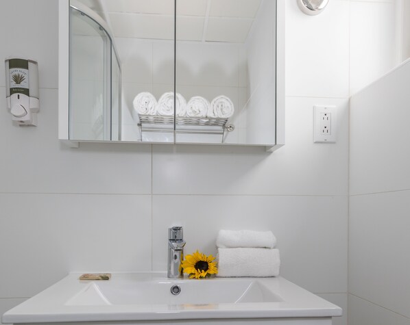 A bathroom with a white sink, a mirror, a towel rack with folded towels, a soap dispenser, and a flower.