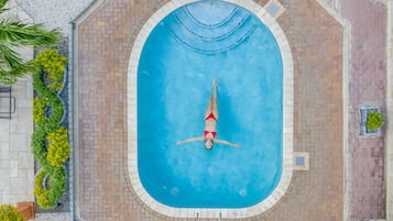 An aerial view of a swimming pool with a person floating in the center, surrounded by a brick deck and greenery.
