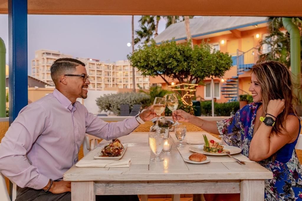 A couple toasting with champagne glasses at an outdoor table with a city view in the background.