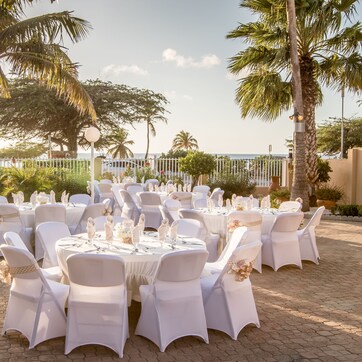 An outdoor wedding setup with round tables and white chairs, surrounded by palm trees and a paved area.