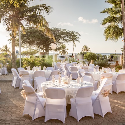 An outdoor wedding setup with round tables and white chairs, surrounded by palm trees and a paved area.