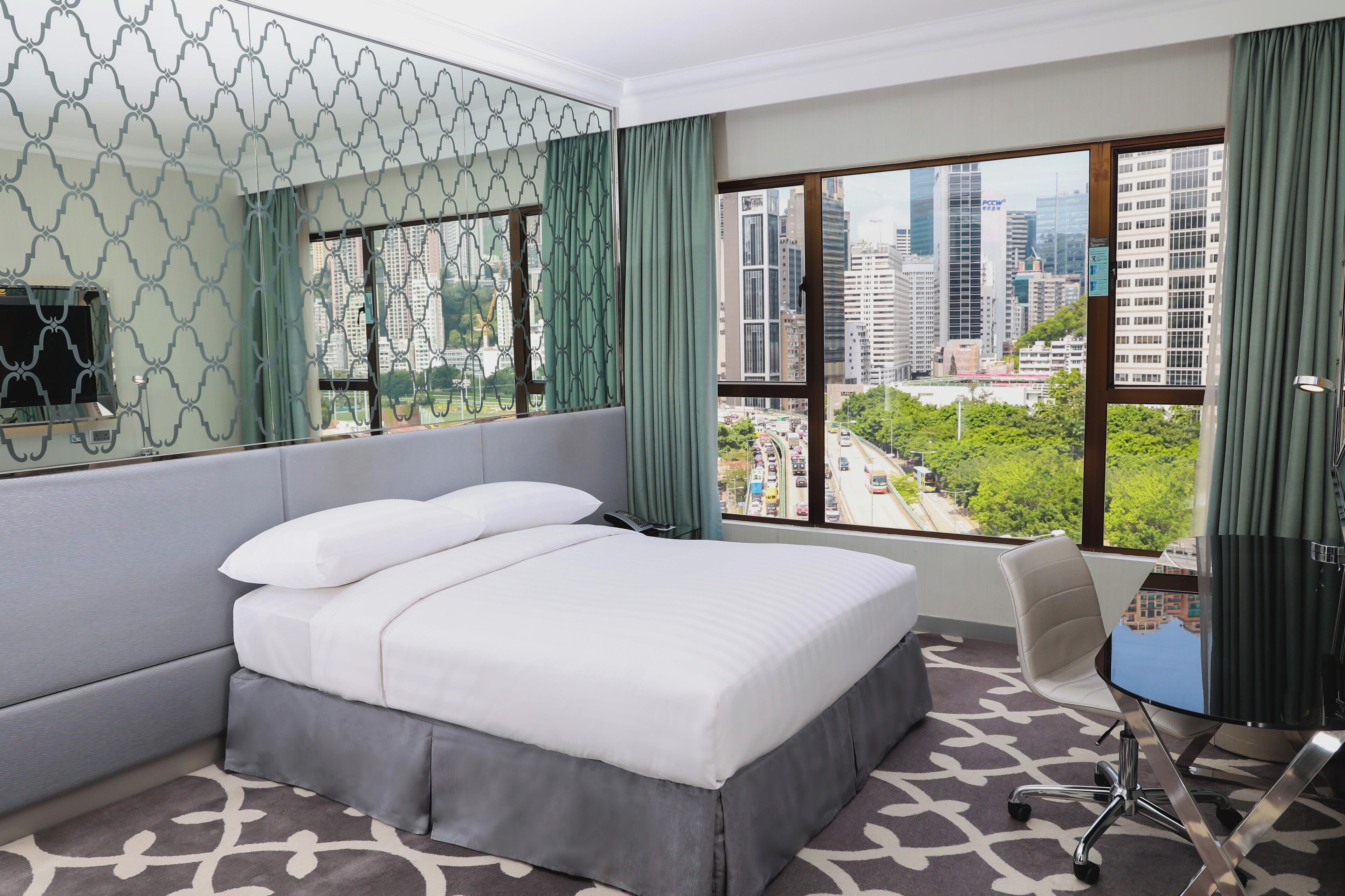 A hotel room with a large bed, a desk with a chair, a view of the cityscape, and a patterned carpet.