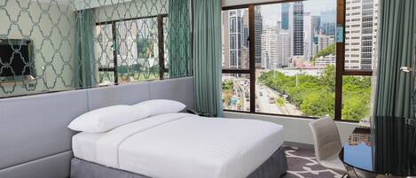 A hotel room with a large bed, a desk with a chair, a view of the cityscape, and a patterned carpet.