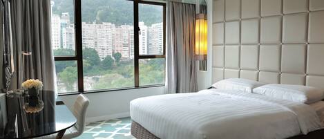 A hotel room with a large bed, a desk with a chair, a view of buildings through the window, and a wall with a padded design.