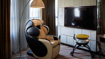 A massage chair, a small table with a yellow object, and a TV in a room with curtains and a patterned carpet.