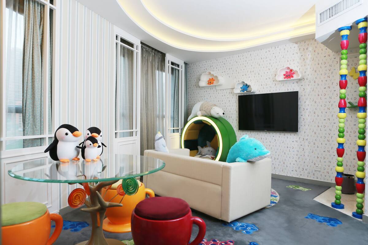 Una sala moderna con una mesa de centro de vidrio, un sofá y un televisor. Hay juguetes de pingüino sobre la mesa y una zona de juegos colorida con un tobogán y una estructura para escalar.