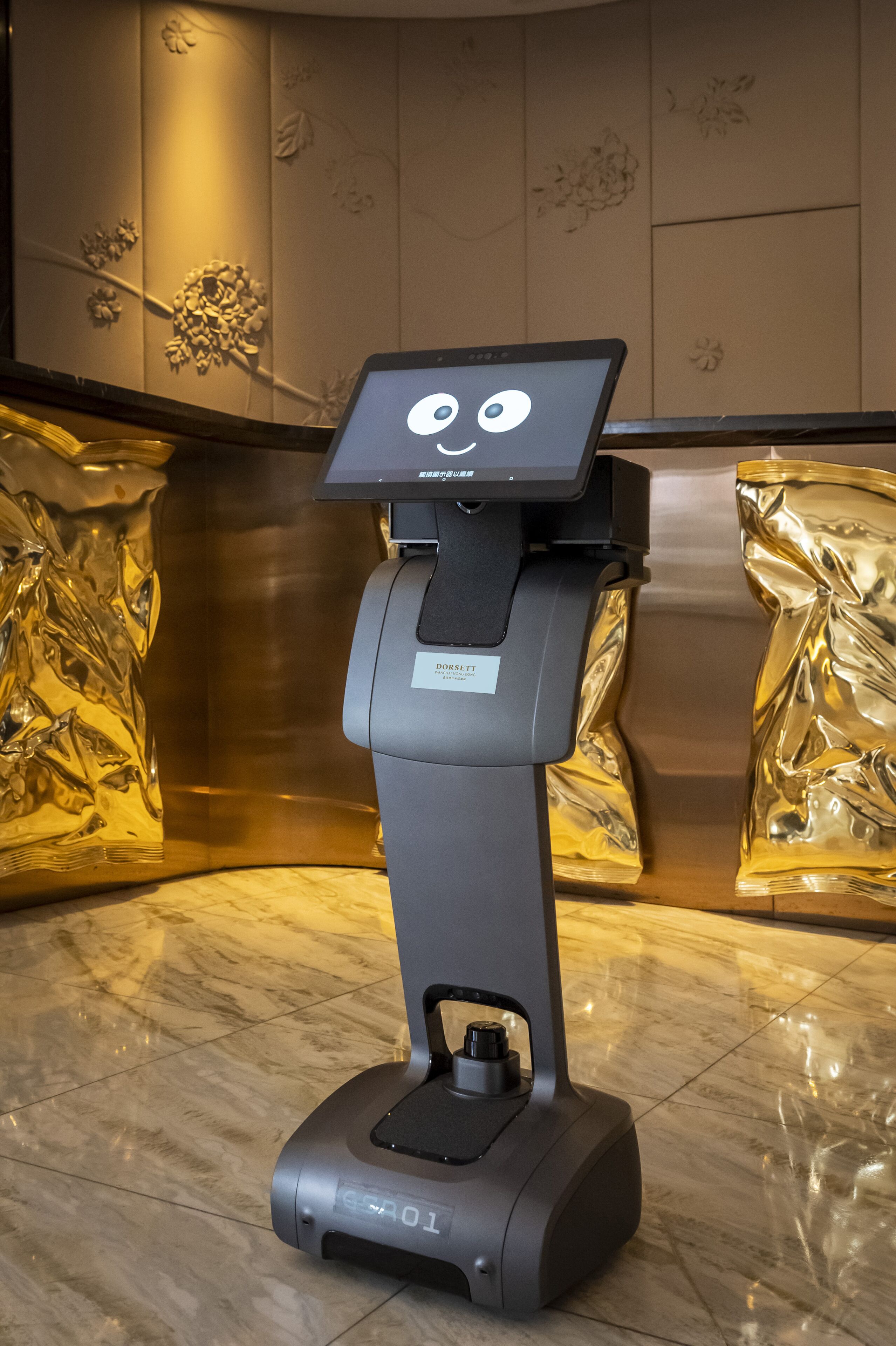 A self-service kiosk with a digital screen displaying a cartoon face, positioned in a lobby with marble flooring and decorative wall panels.