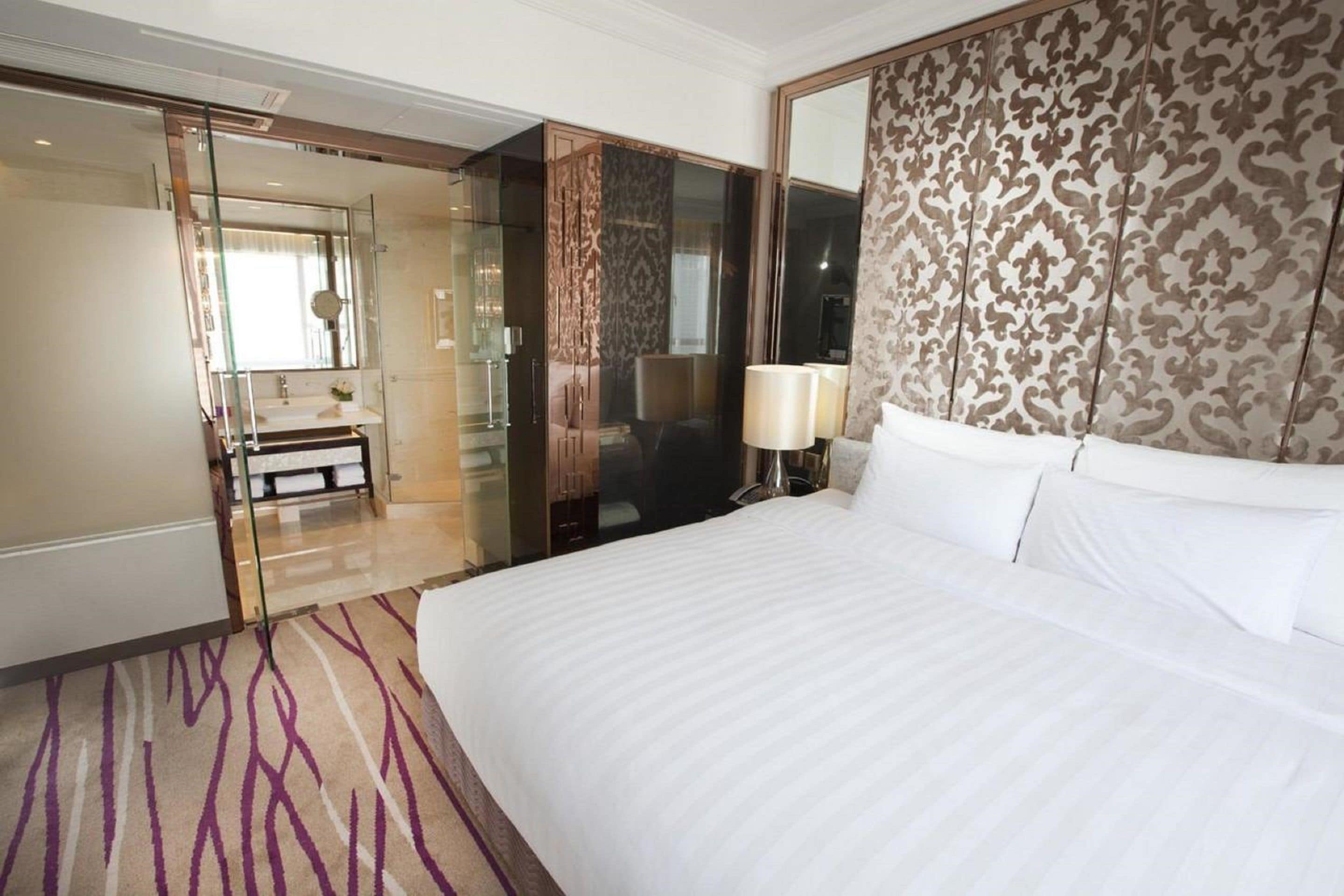 Habitación de hotel con una cama grande, cabina de ducha de vidrio, tocador con espejo y cabecero estampado.