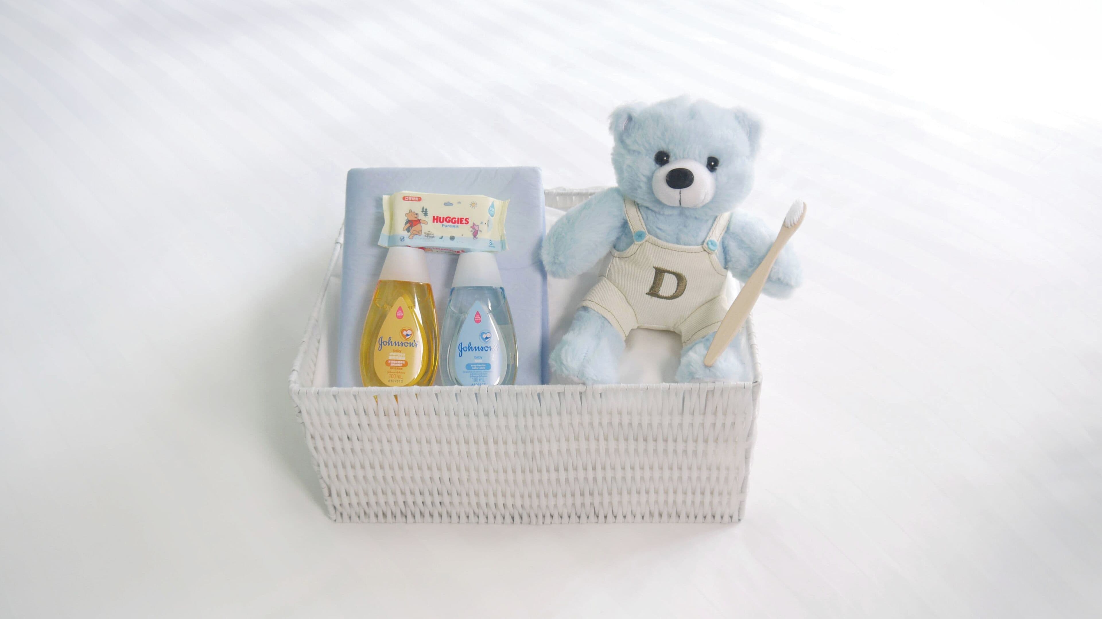 Una cesta blanca de mimbre con productos para bebés, como pañales, toallitas húmedas y un osito de peluche azul.