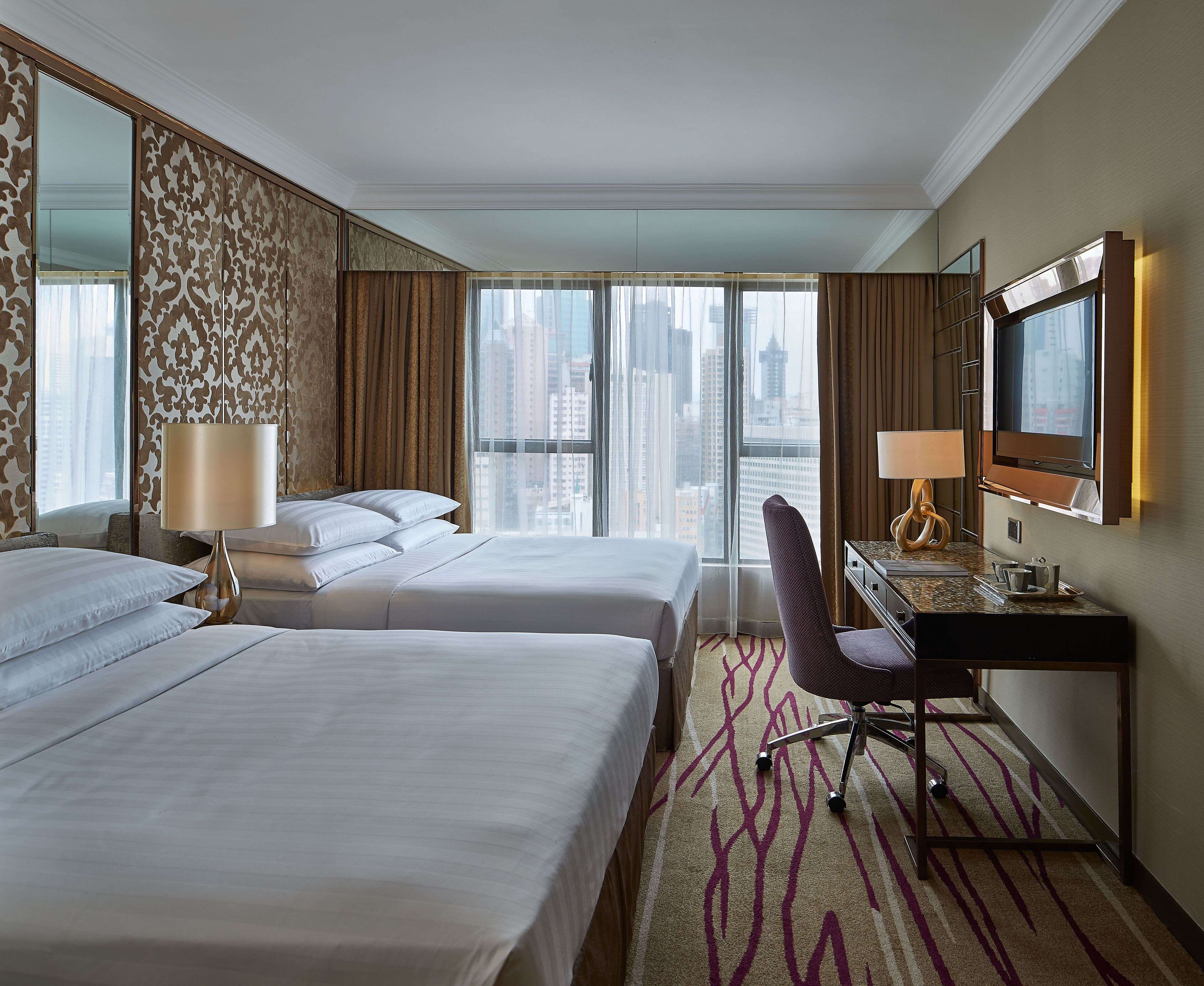 A hotel room with two beds, a desk with a chair, a large mirror, and a view of the cityscape through the window.