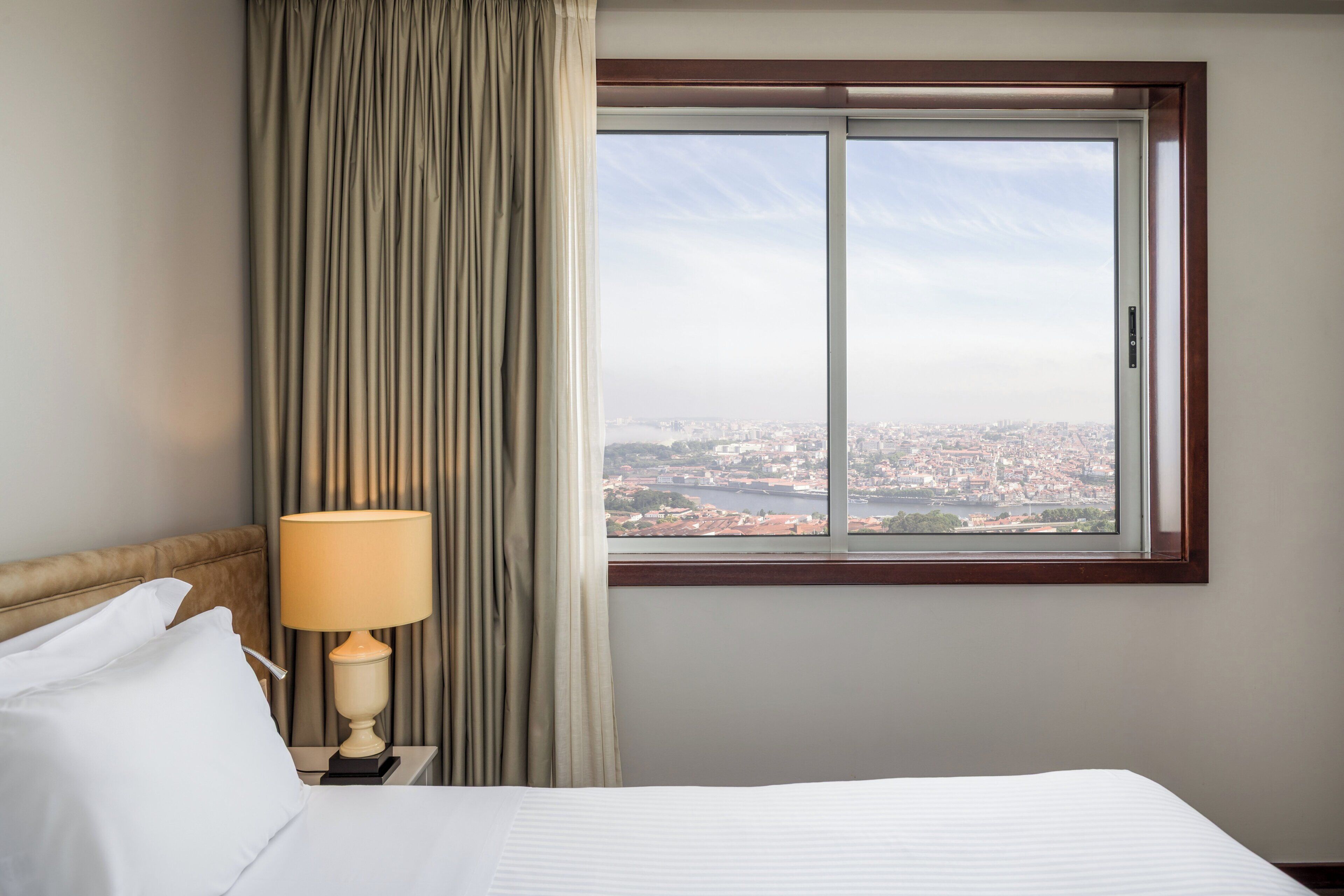 Habitación de hotel con cama, mesita de noche, lámpara, ventana y vistas a la ciudad, además de cortinas.