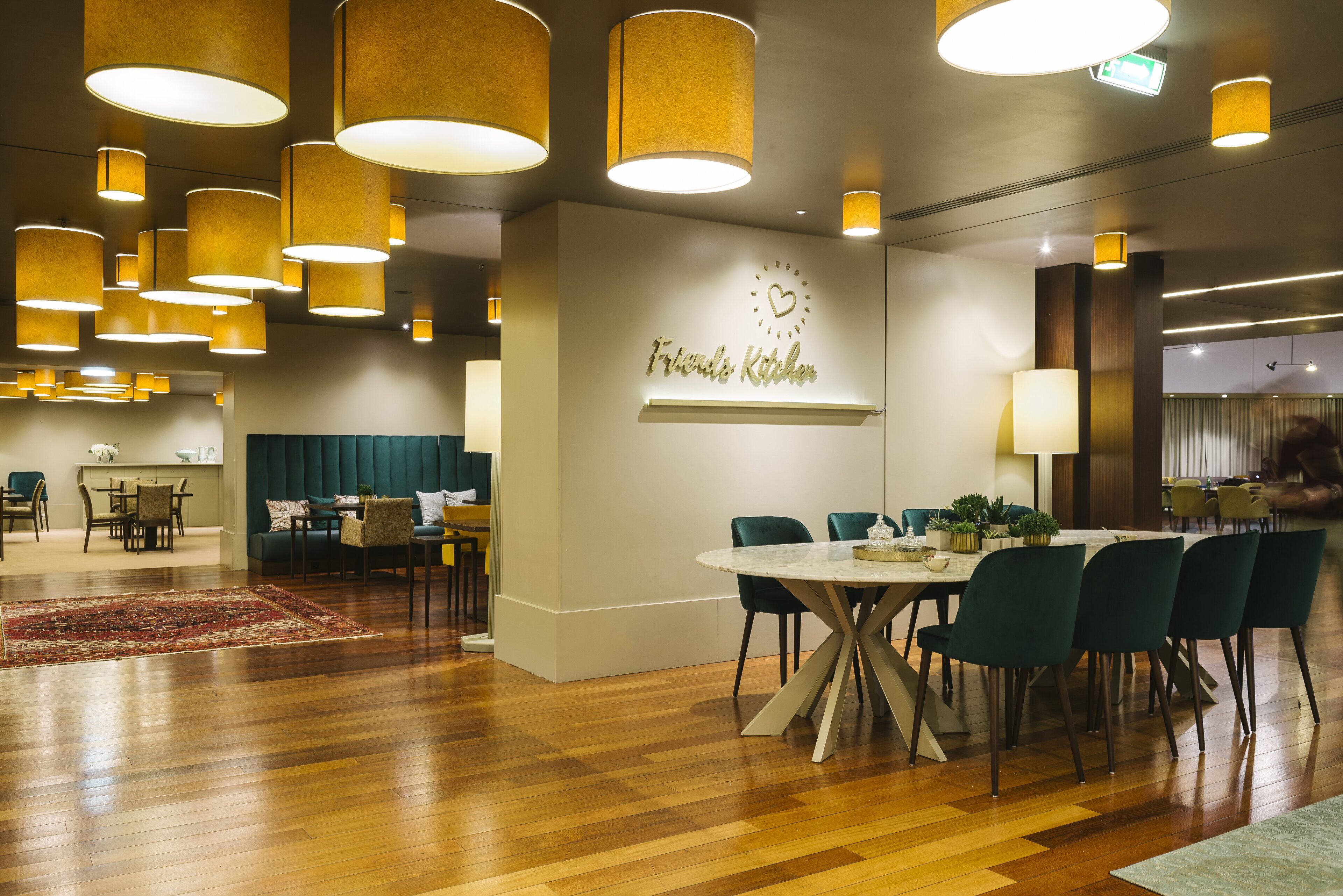 A modern dining area with a round table, green chairs, and a 'Friends Kitchen' sign on the wall.