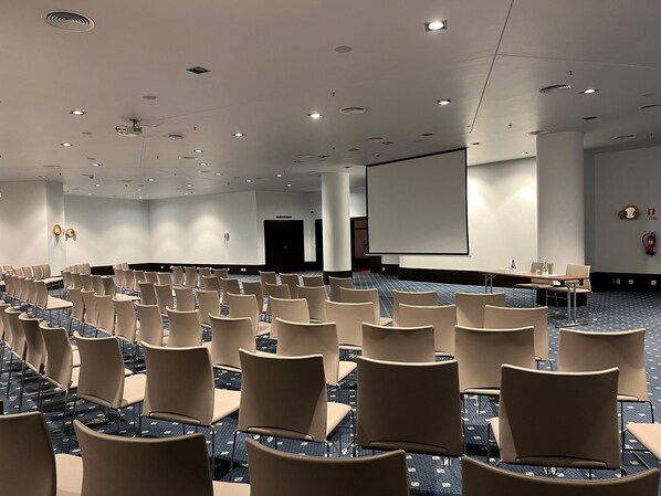 Un salón de conferencias con filas de sillas, una pantalla grande y un proyector.