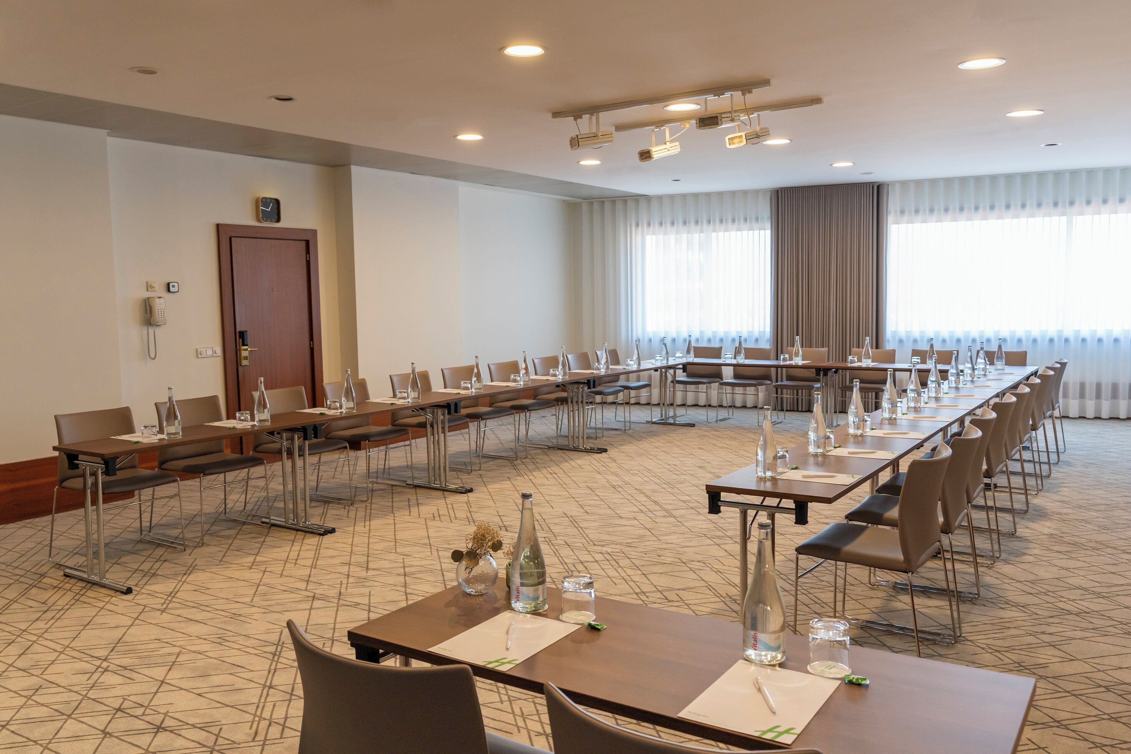 Une salle de conférence dotée de plusieurs tables et de chaises, équipée de bouteilles d’eau et de verres, d’une horloge au mur et d’une porte.