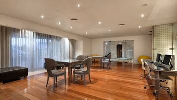Quarto de hotel moderno com piso de madeira, área de refeições com cadeiras e mesa, cama com divisória de vidro e escrivaninha com computador.