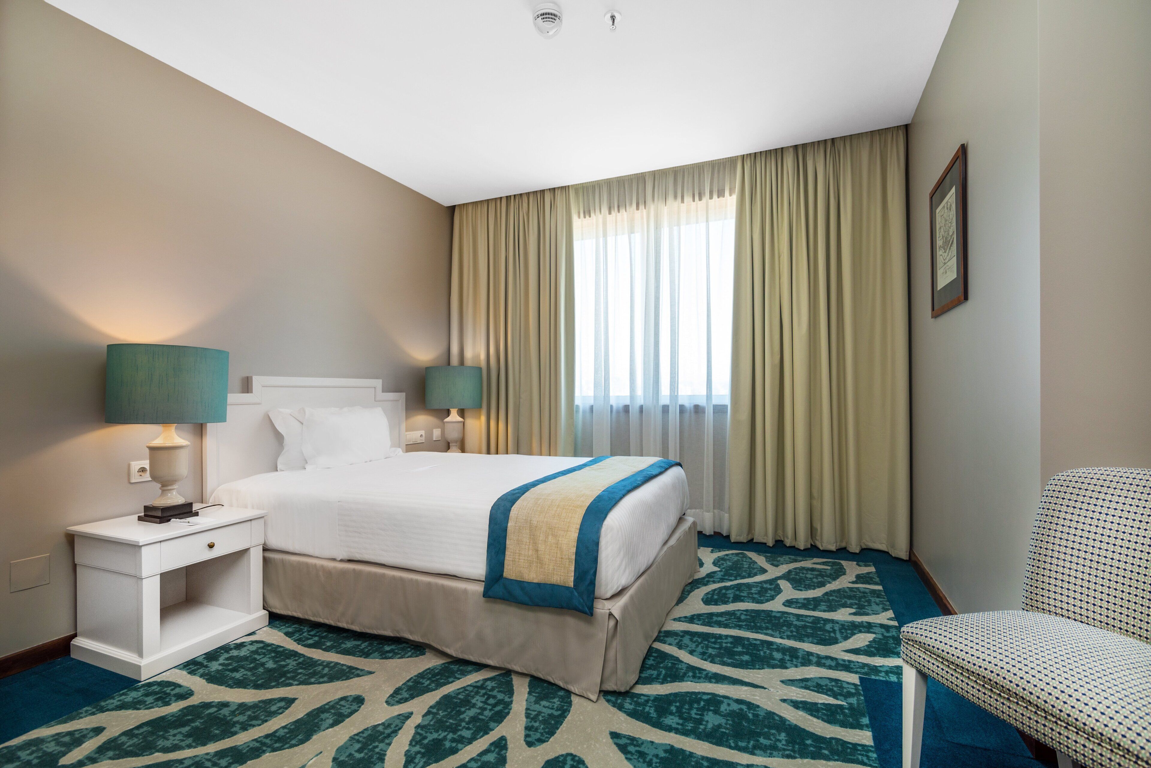 A hotel room with a bed, two bedside tables, a chair, a window with curtains, and a carpet with a leaf pattern.