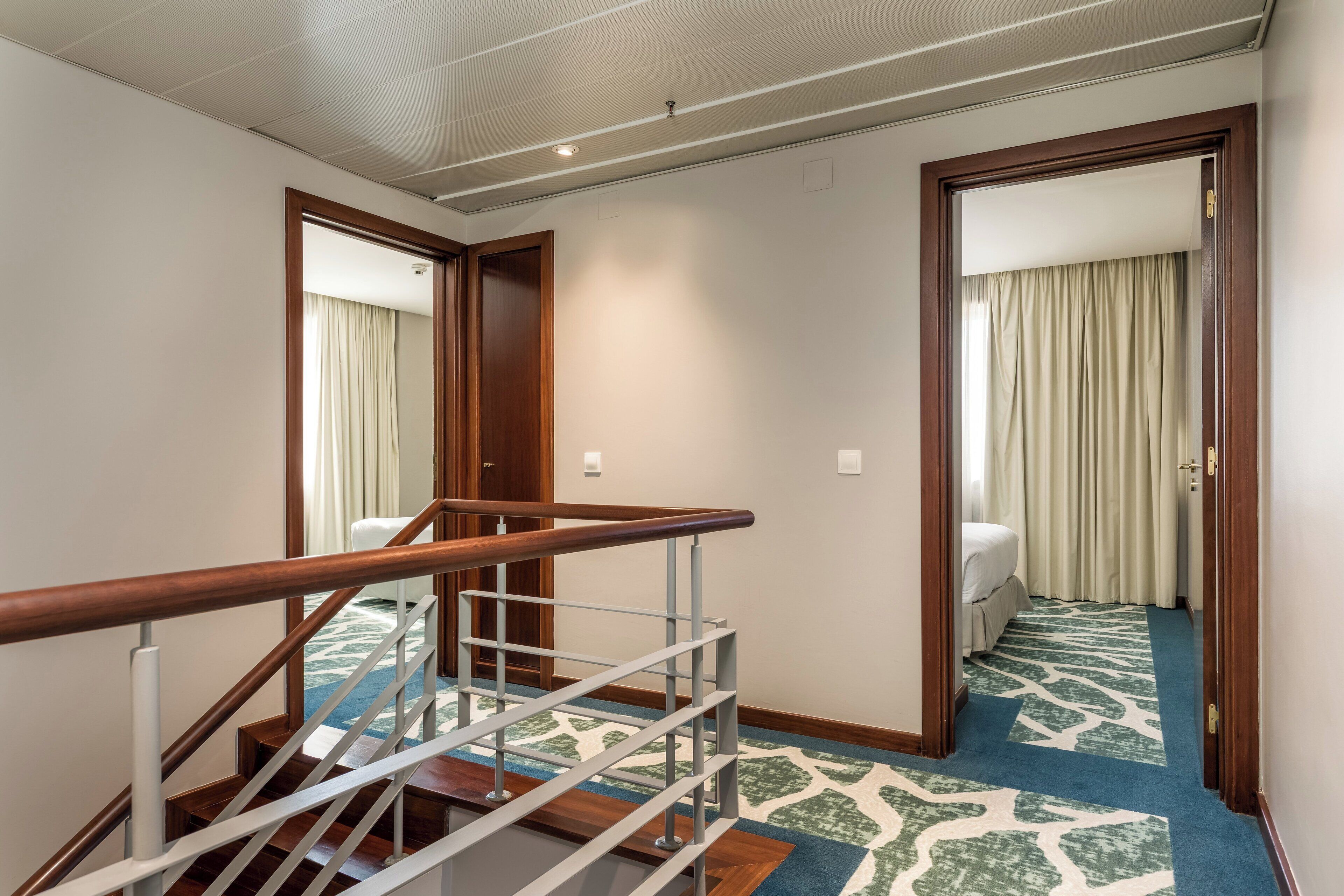 Een hotelgangen met een houten trap, een kamerbreed tapijt en een bed in de aangrenzende kamer.