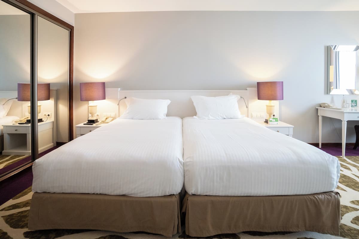 Un lit double avec du linge de lit blanc, deux tables de chevet avec des lampes, un bureau avec un miroir et un grand miroir fixé au mur.