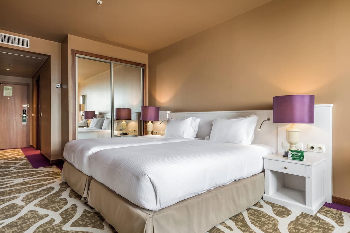 Une chambre d’hôtel avec un grand lit, une table de chevet avec une lampe, un miroir et un sol recouvert d’une moquette.