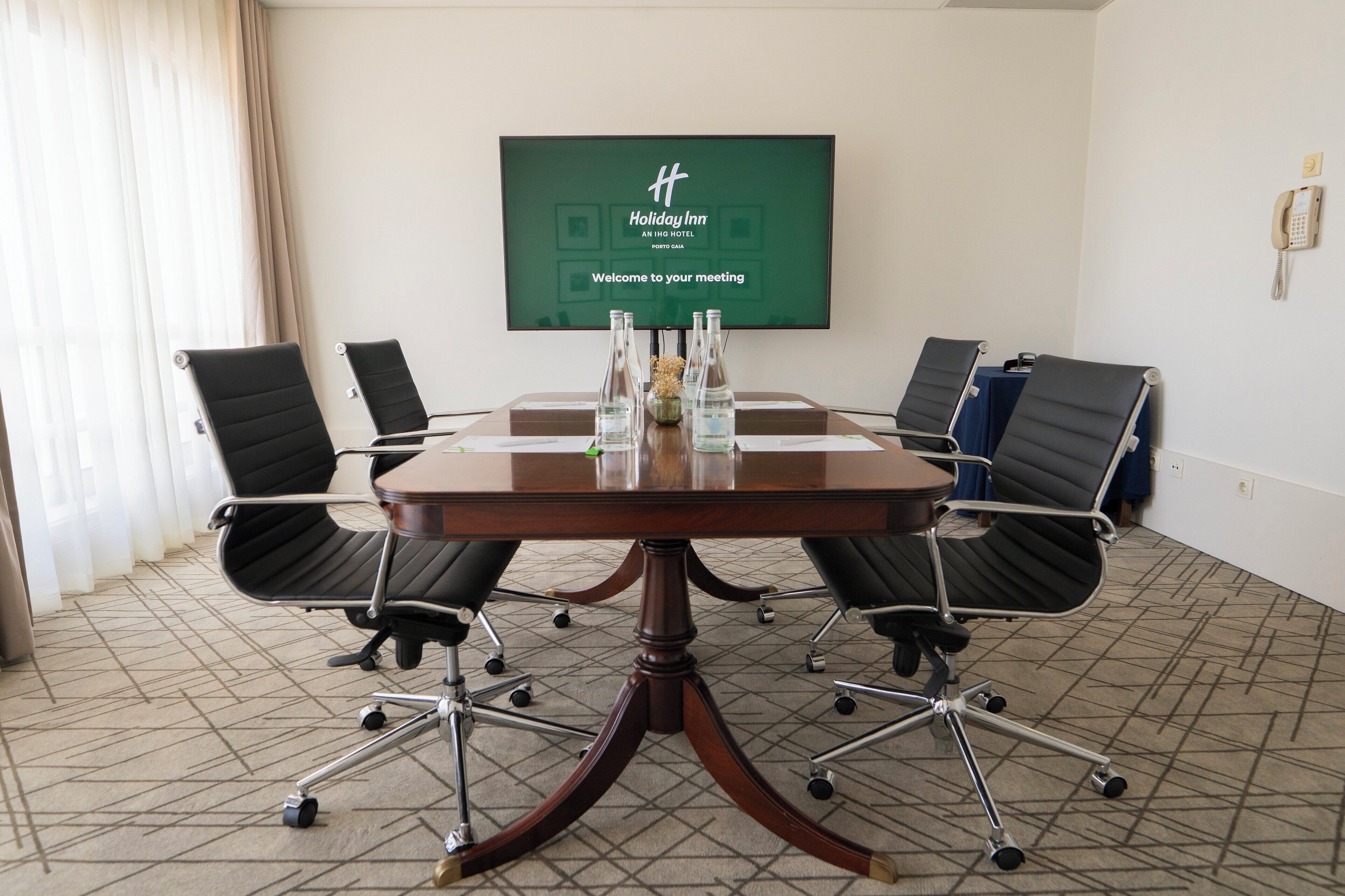 A conference room with a round table, four black office chairs, and a screen displaying "Holiday Inn Anling Hotel, Porto Dae Welcome to your meeting".