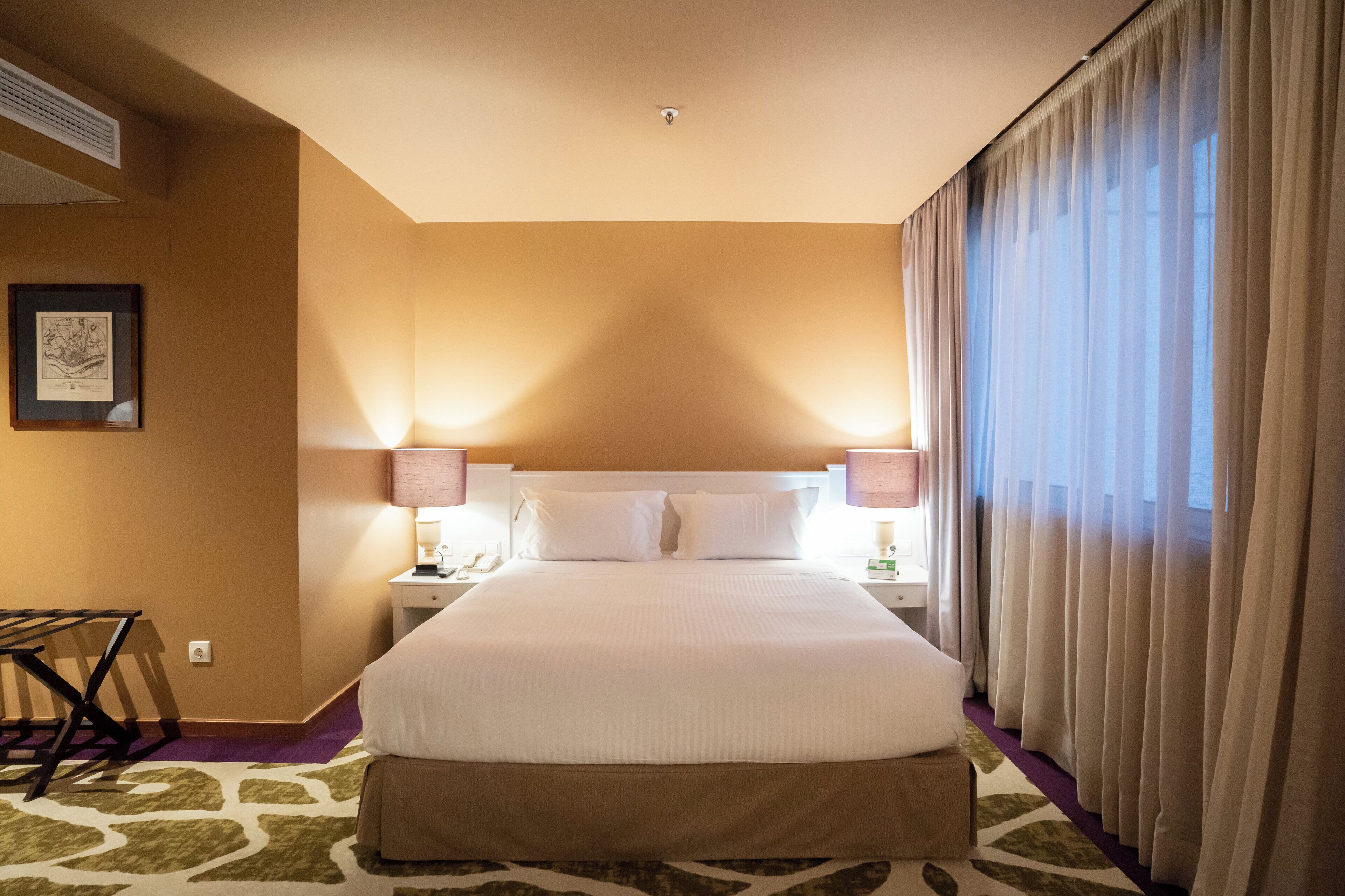 Habitación de hotel con una cama grande, dos lámparas de noche, una mesita de noche, un cuadro en la pared y una ventana con cortinas.