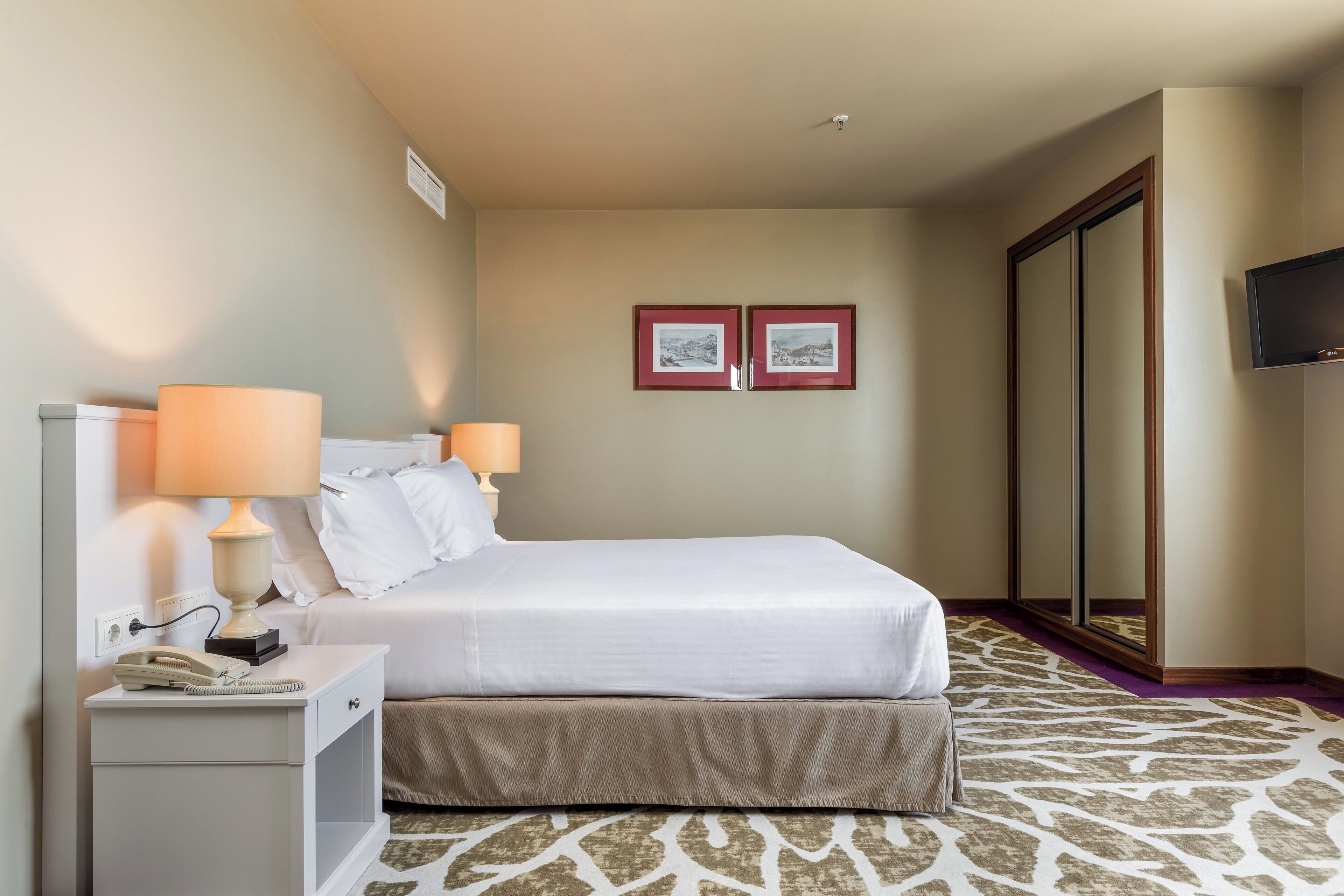 A hotel room with a bed, bedside lamps, a telephone, a mirror, and two framed pictures on the wall.