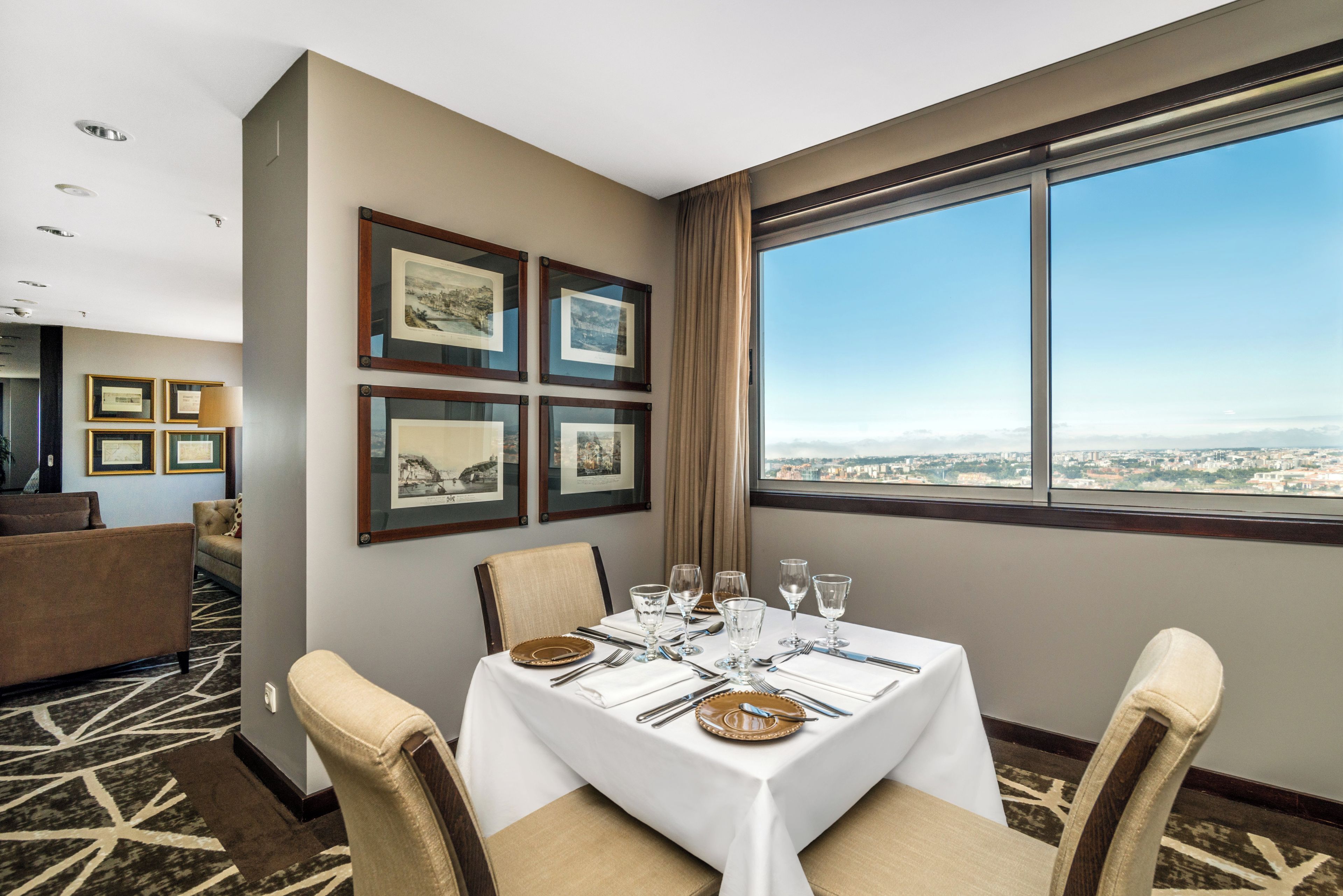 Un comedor de hotel con una mesa preparada para cuatro personas, fotografías enmarcadas en la pared y vistas de un paisaje urbano a través de un amplio ventanal.
