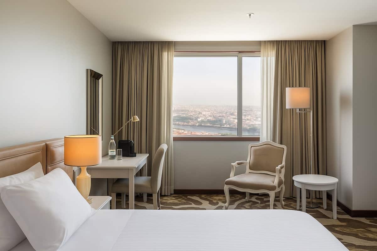 Une chambre d’hôtel équipée d’un lit, d’un bureau, d’une chaise, d’une petite table, d’une lampe et offrant une vue sur la ville grâce à une grande fenêtre.