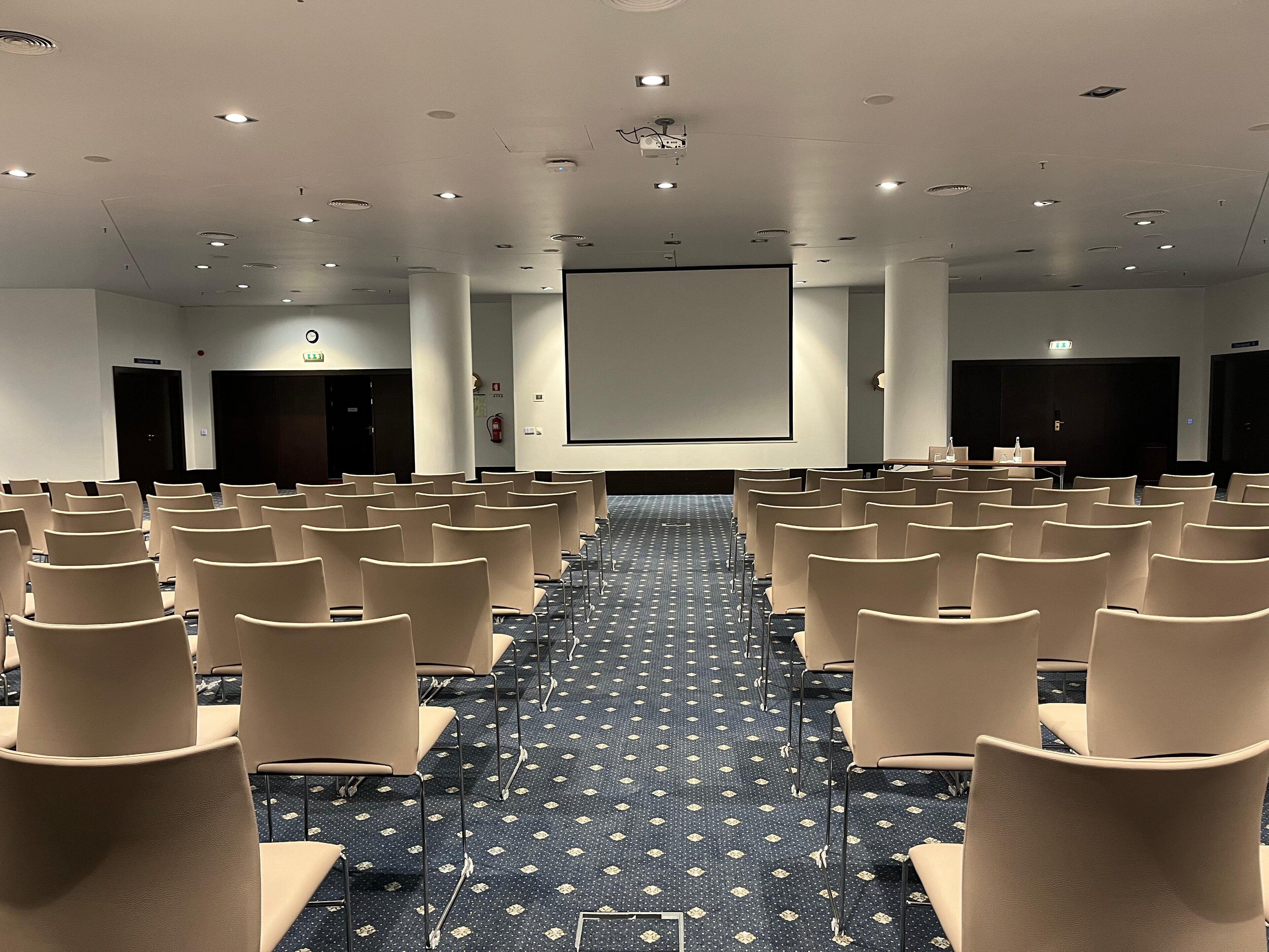 Une salle de conférence avec des rangées de chaises beiges, un grand écran à l’avant et un tapis à motifs.