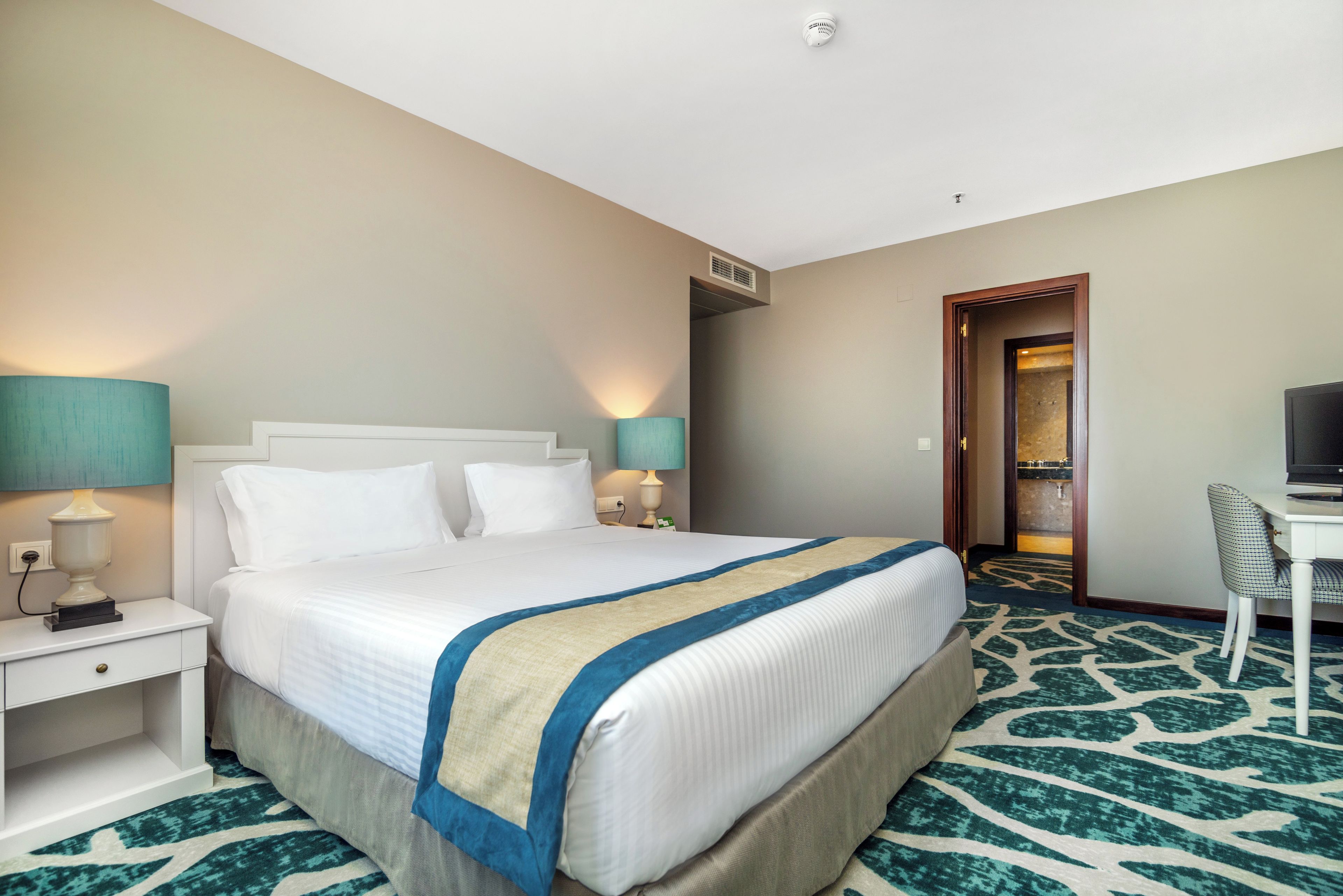 A hotel room with a bed, bedside tables, a desk, a chair, and a television.