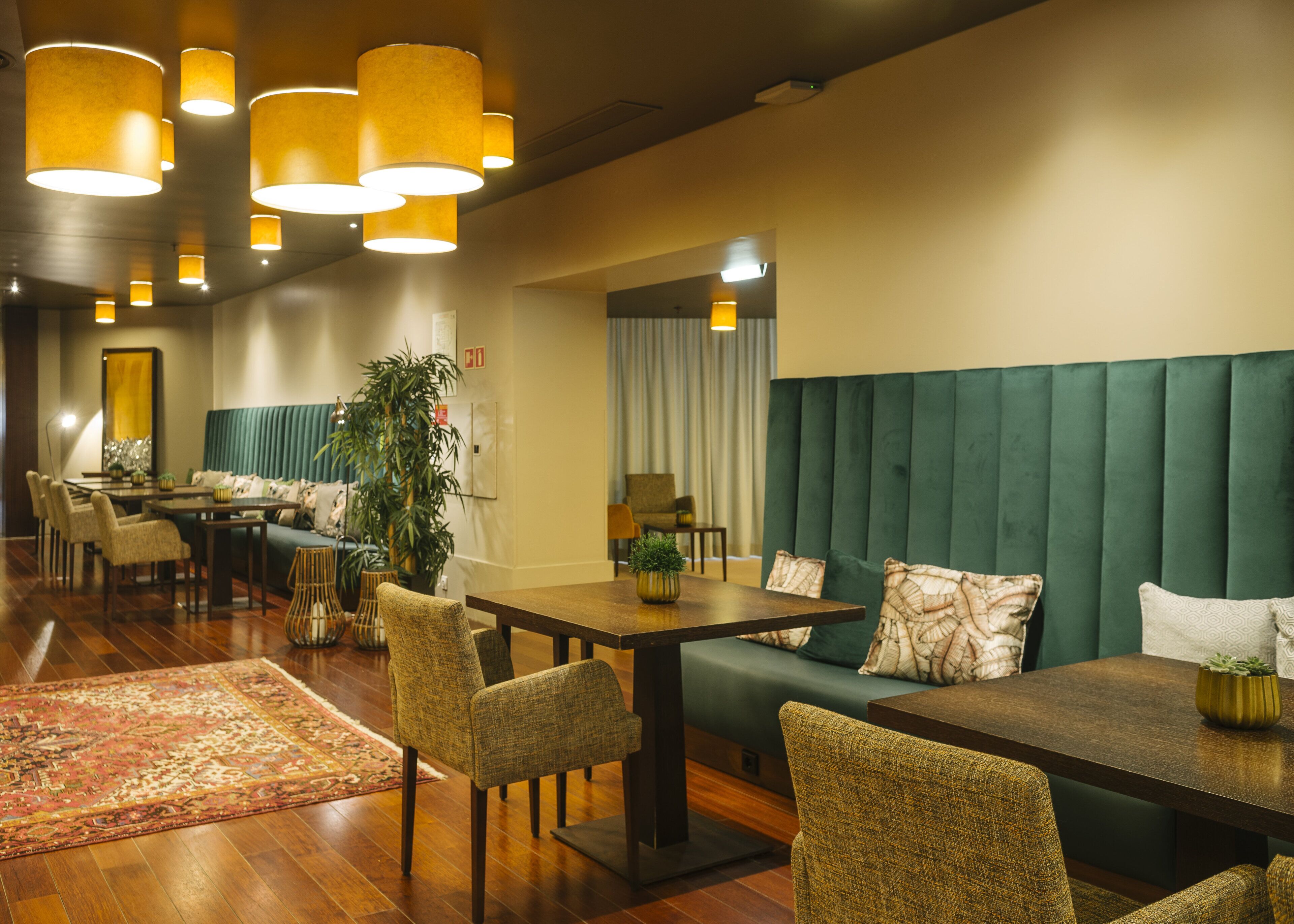 Un moderno vestíbulo de hotel con un sofá tapizado en verde, mesas y sillas de madera. Se observan varias lámparas colgantes y una planta grande en maceta.